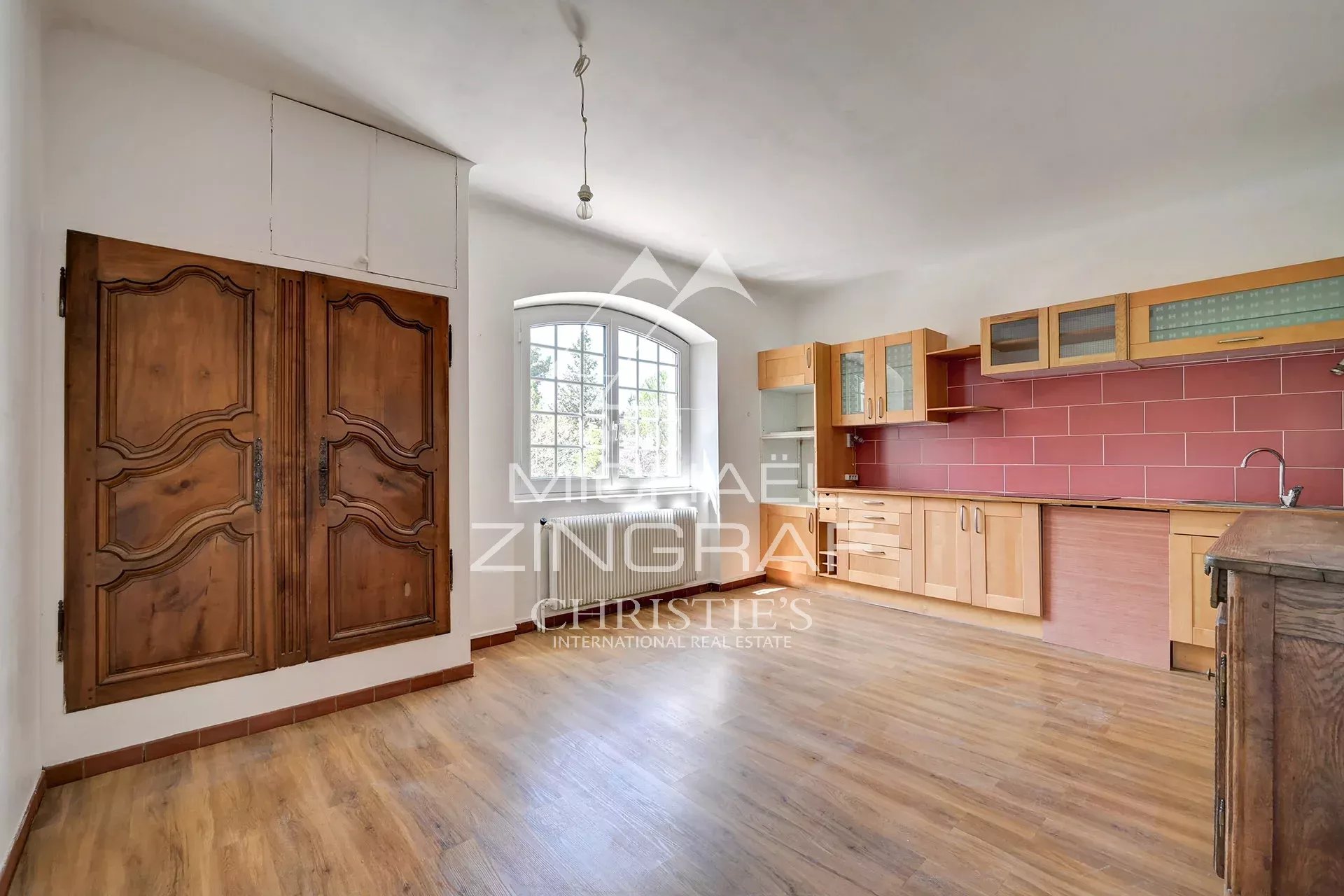 Rustic kitchen with wooden cabinets and a red tile backsplash, arched window, and large carved doors on the left.