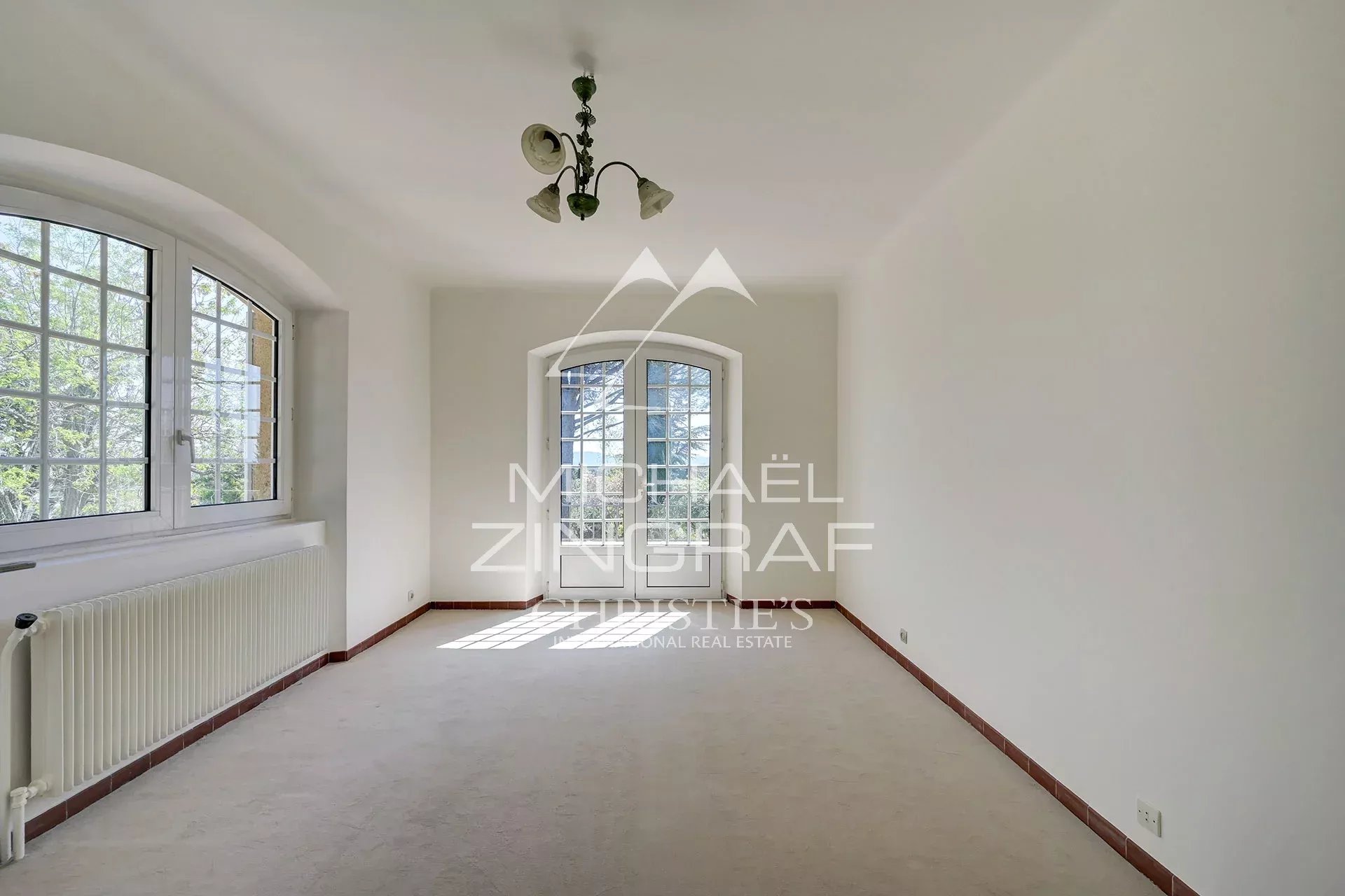 Bright, empty living room with white walls, a curved arched window/door at the far end, and a radiator along the left wall, lit by natural daylight.