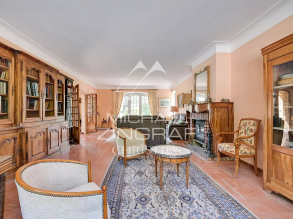 Traditional living room with peach walls, a long wooden bookcase wall on the left, and a brick fireplace on the right. A seating area with sofas and armchairs sits on a patterned rug in front of a large window.