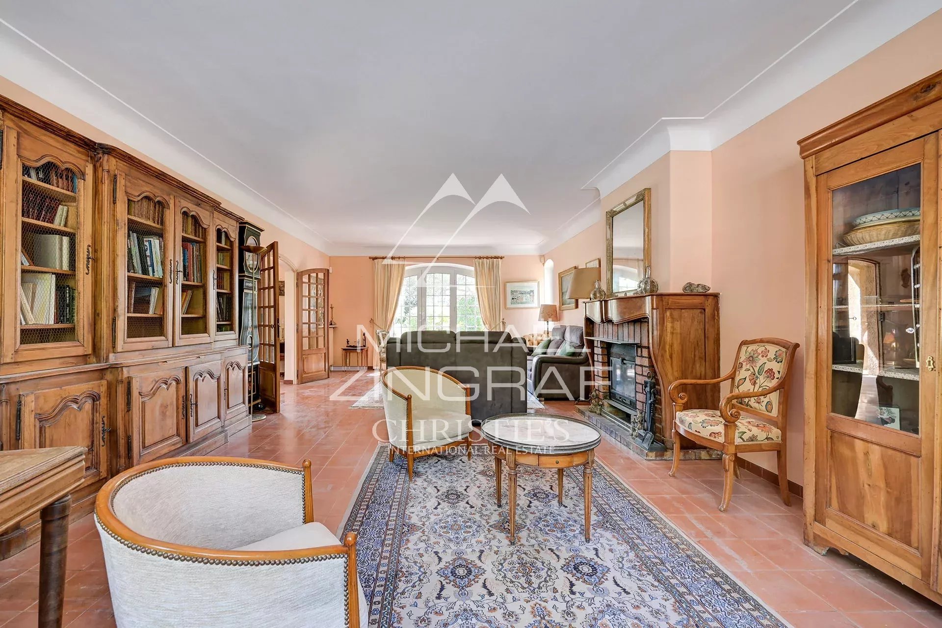 Traditional living room with peach walls, a long wooden bookcase wall on the left, and a brick fireplace on the right. A seating area with sofas and armchairs sits on a patterned rug in front of a large window.