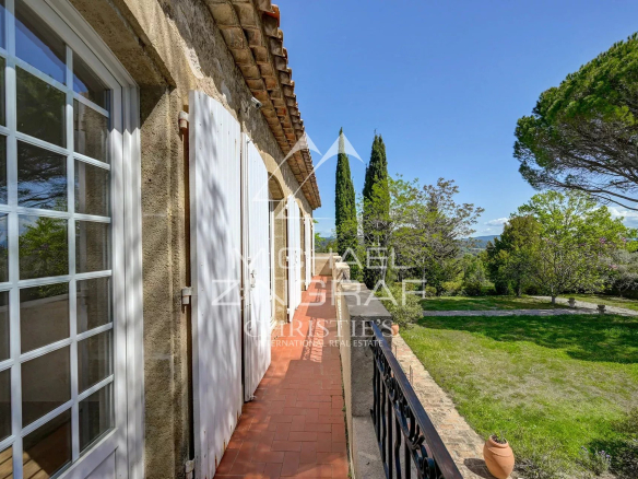 Terracotta-tiled balcony of a stone house with white shutters, overlooking a green garden and blue sky.