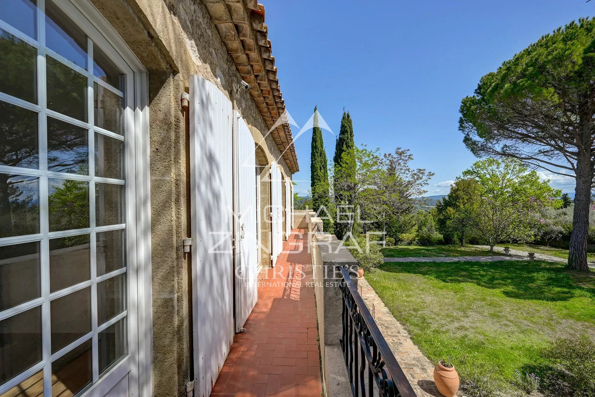 Terracotta-tiled balcony of a stone house with white shutters, overlooking a green garden and blue sky.