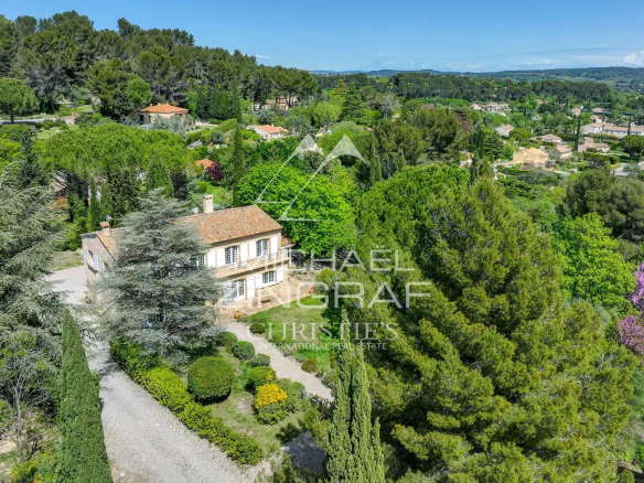 A stone house with a red-tiled roof surrounded by manicured gardens and tall trees in a green hillside neighborhood.