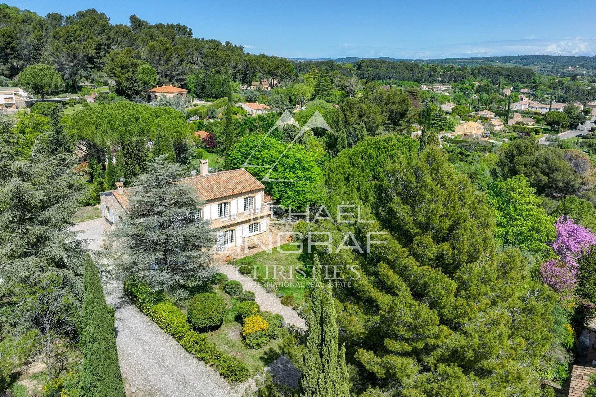 A stone house with a red-tiled roof surrounded by manicured gardens and tall trees in a green hillside neighborhood.