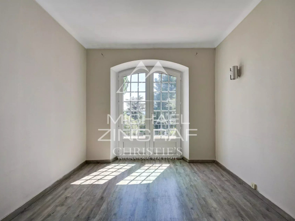 Empty room with arched French doors opening to a garden; beige walls and gray wood flooring, sunlight on the floor.
