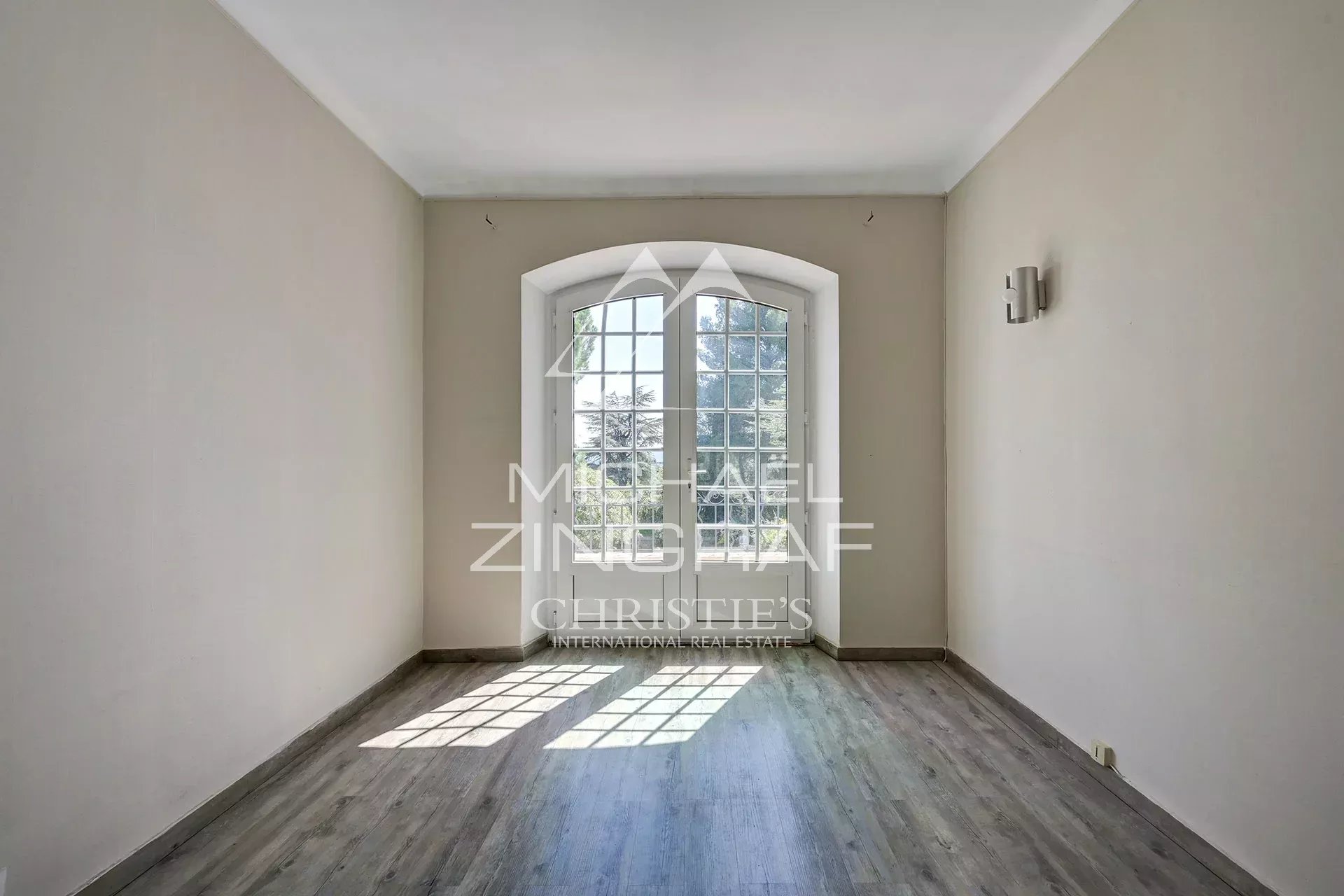 Empty room with arched French doors opening to a garden; beige walls and gray wood flooring, sunlight on the floor.
