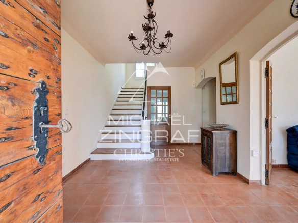 Bright entry hall with terracotta tiled floor, wooden front door to the left, and a staircase under a wrought‑iron chandelier.