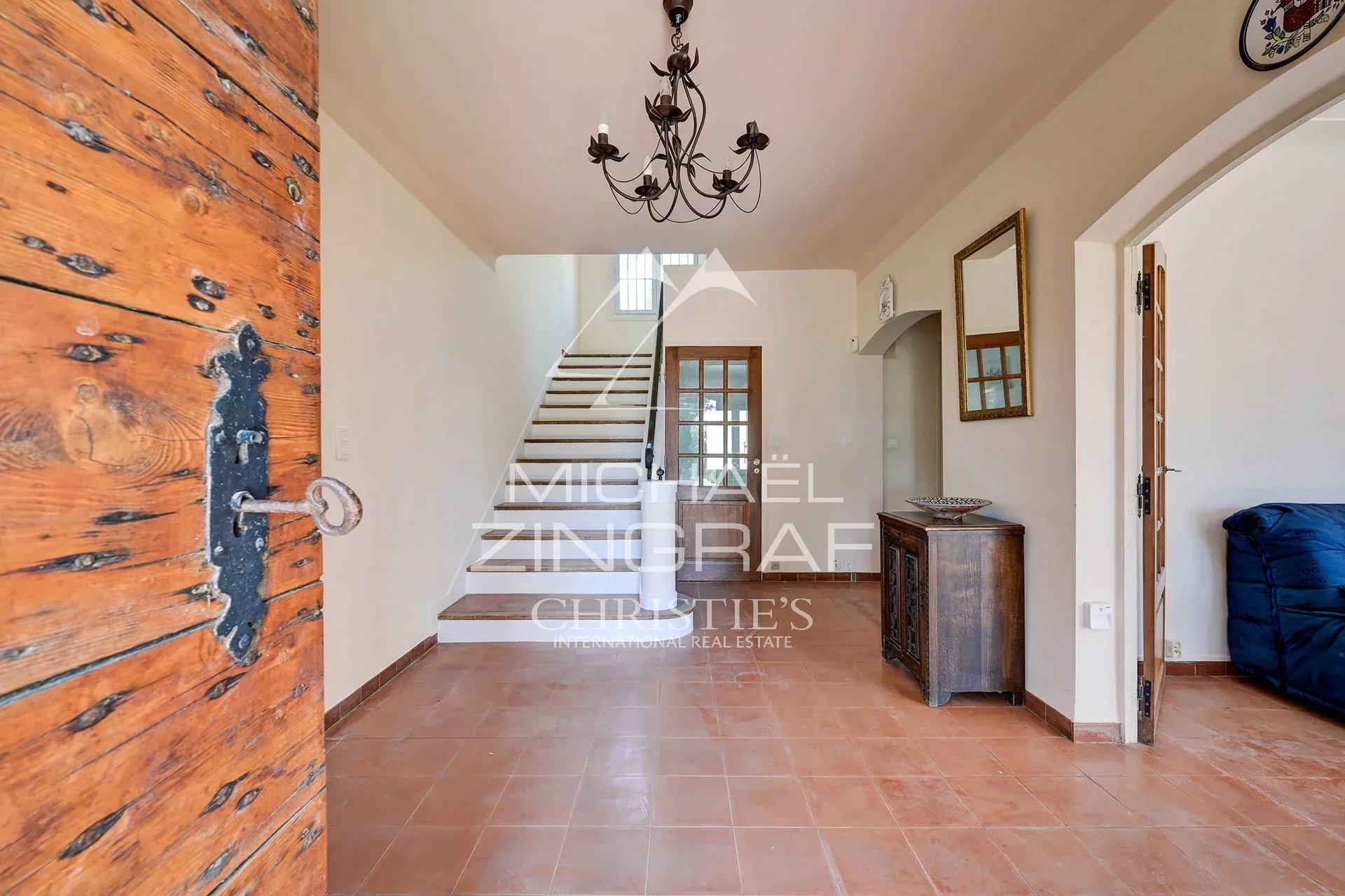 Bright entry hall with terracotta tiled floor, wooden front door to the left, and a staircase under a wrought‑iron chandelier.