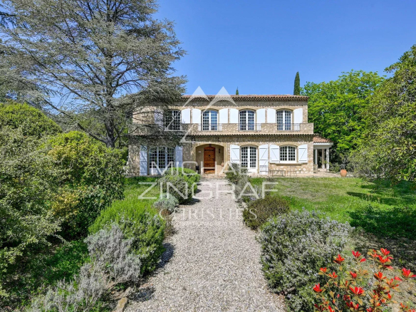Two-story stone villa with white shutters, arched windows, and a gravel path leading to the wooden front door amid a lush garden.