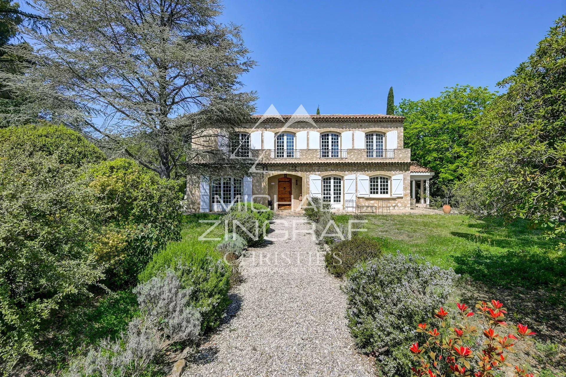 Two-story stone villa with white shutters, arched windows, and a gravel path leading to the wooden front door amid a lush garden.