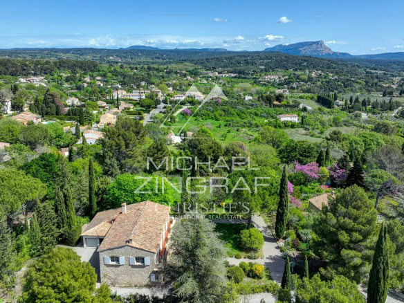 Aerial view of a green, rural village with trees, shrubs, and scattered houses under a blue sky and distant mountains.