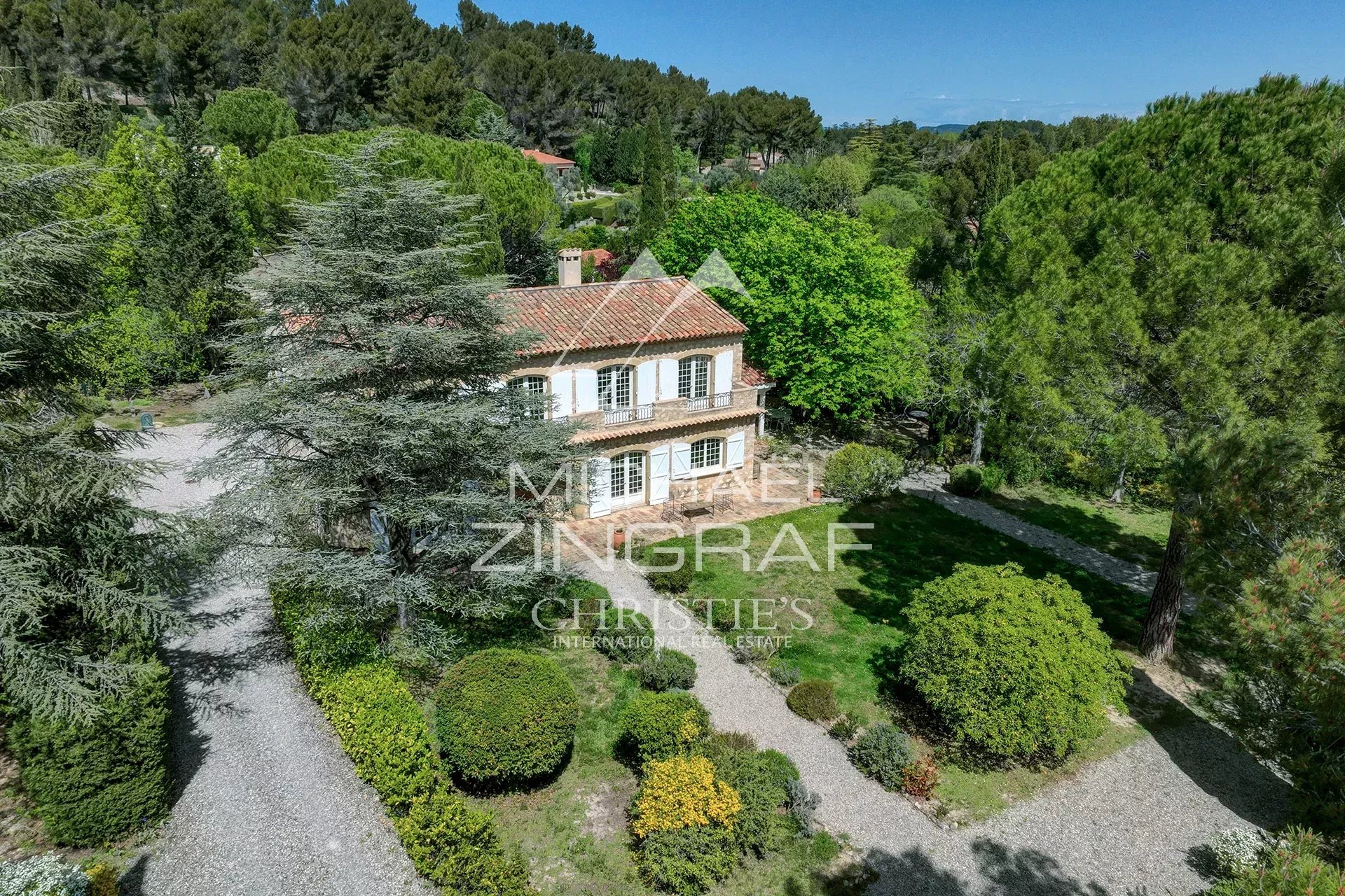 Two-story stone villa with a terracotta roof, white shutters, and a manicured gravel driveway tucked among tall trees and lush greenery outside. (Property listing)