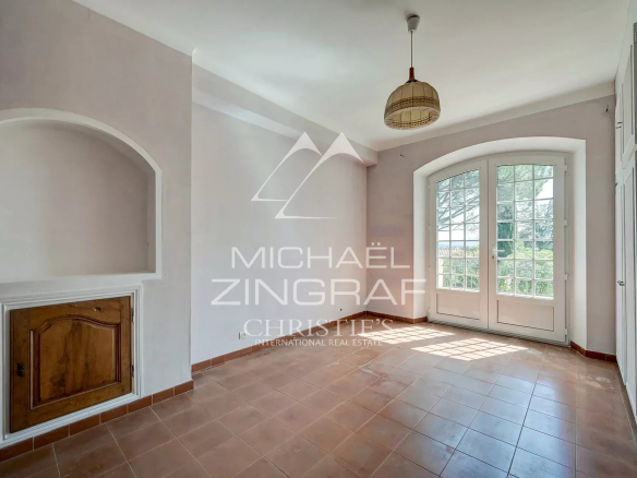 Bright empty room with terracotta tile floor, white walls, an arched alcove with a wooden cabinet, and double French doors opening to outside.