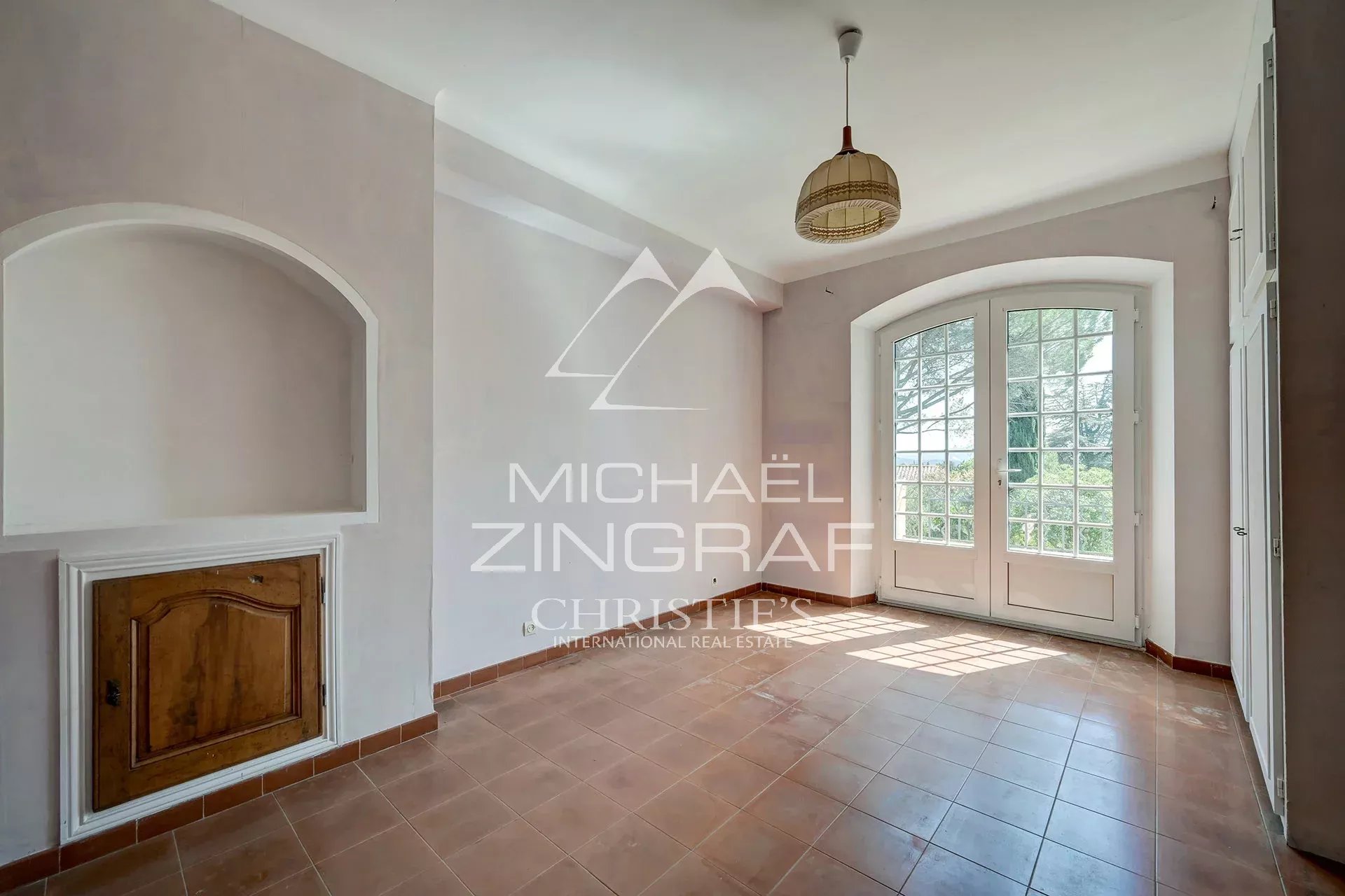 Bright empty room with terracotta tile floor, white walls, an arched alcove with a wooden cabinet, and double French doors opening to outside.