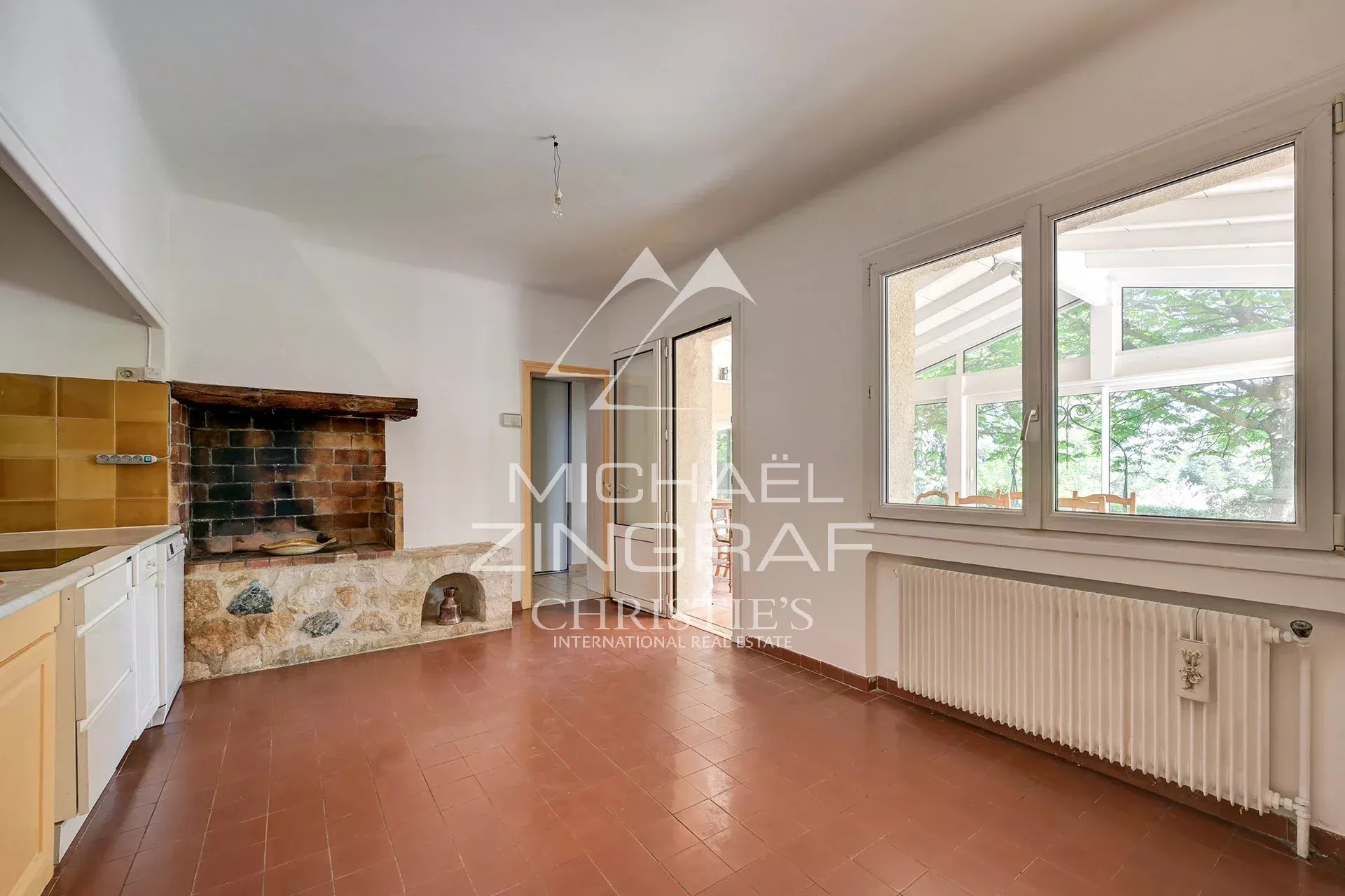 Bright kitchen featuring a rustic brick fireplace with stone base, white cabinetry, and terracotta tiles; large windows overlook a green conservatory outside.