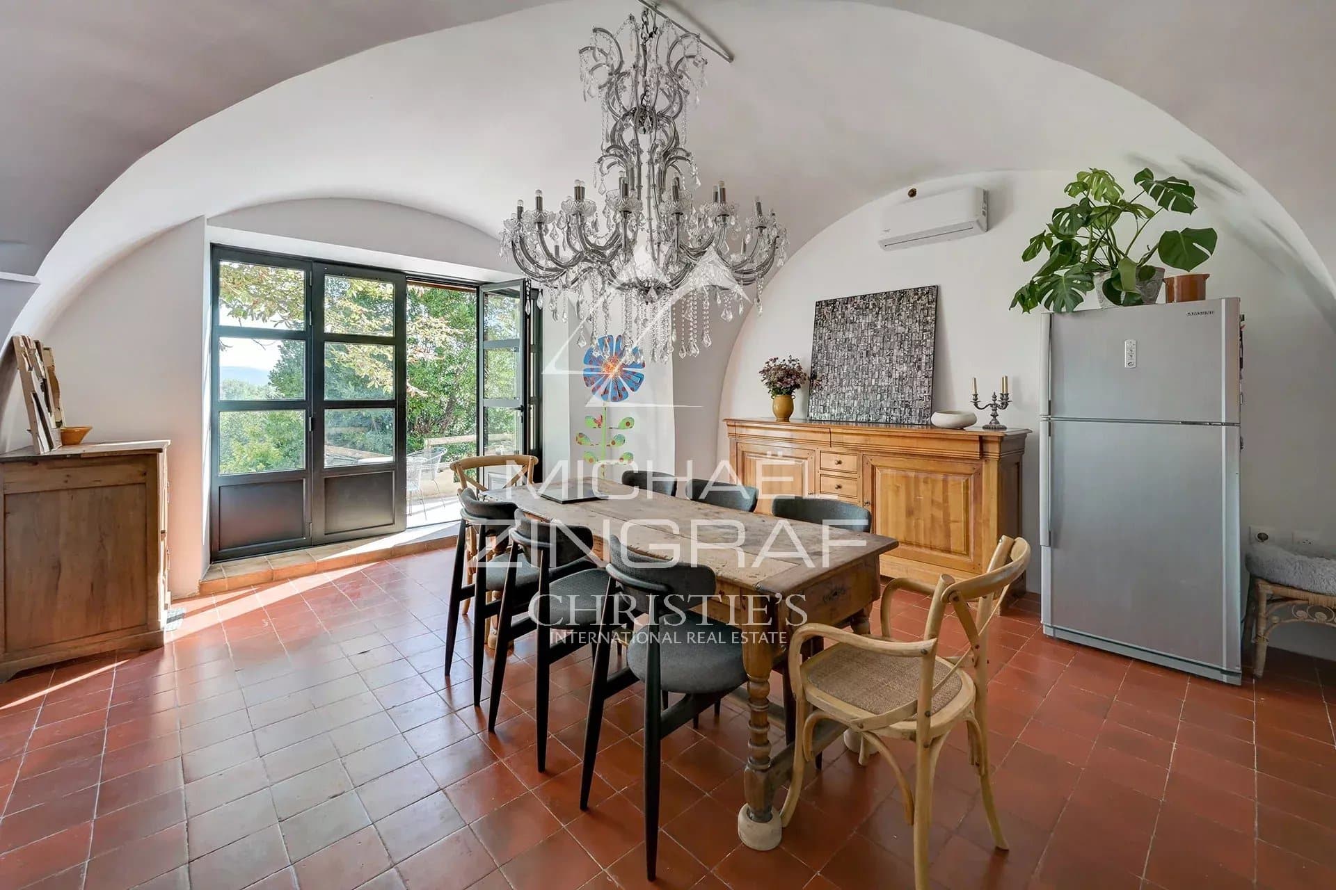Intérieur d'une maison avec une table en bois, des chaises, et une grande fenêtre donnant sur un paysage.