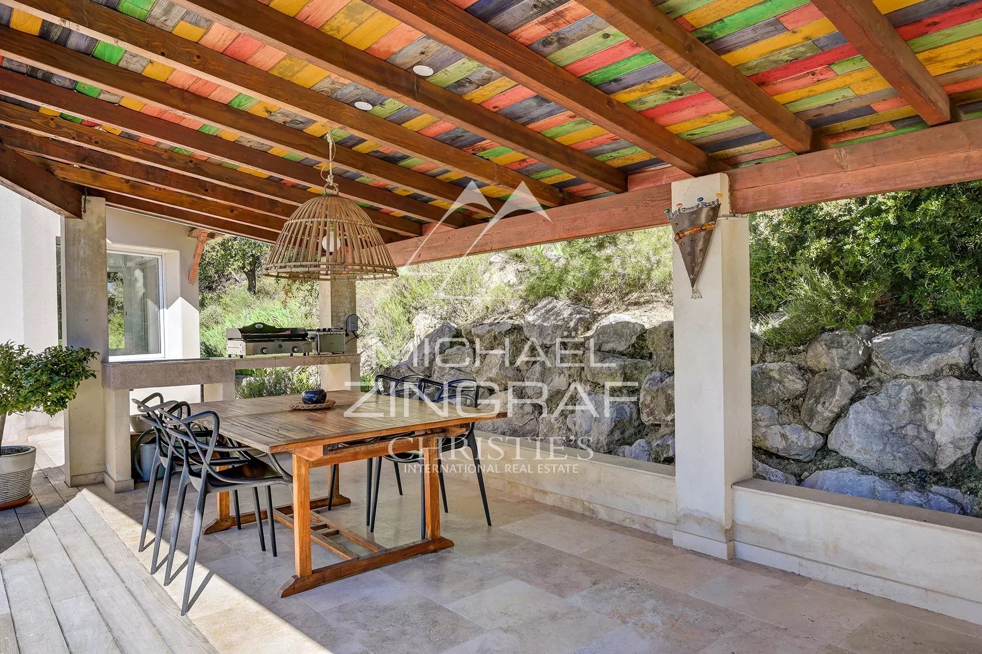 Wooden terrace with table and chairs under a colorful awning, surrounded by greenery.