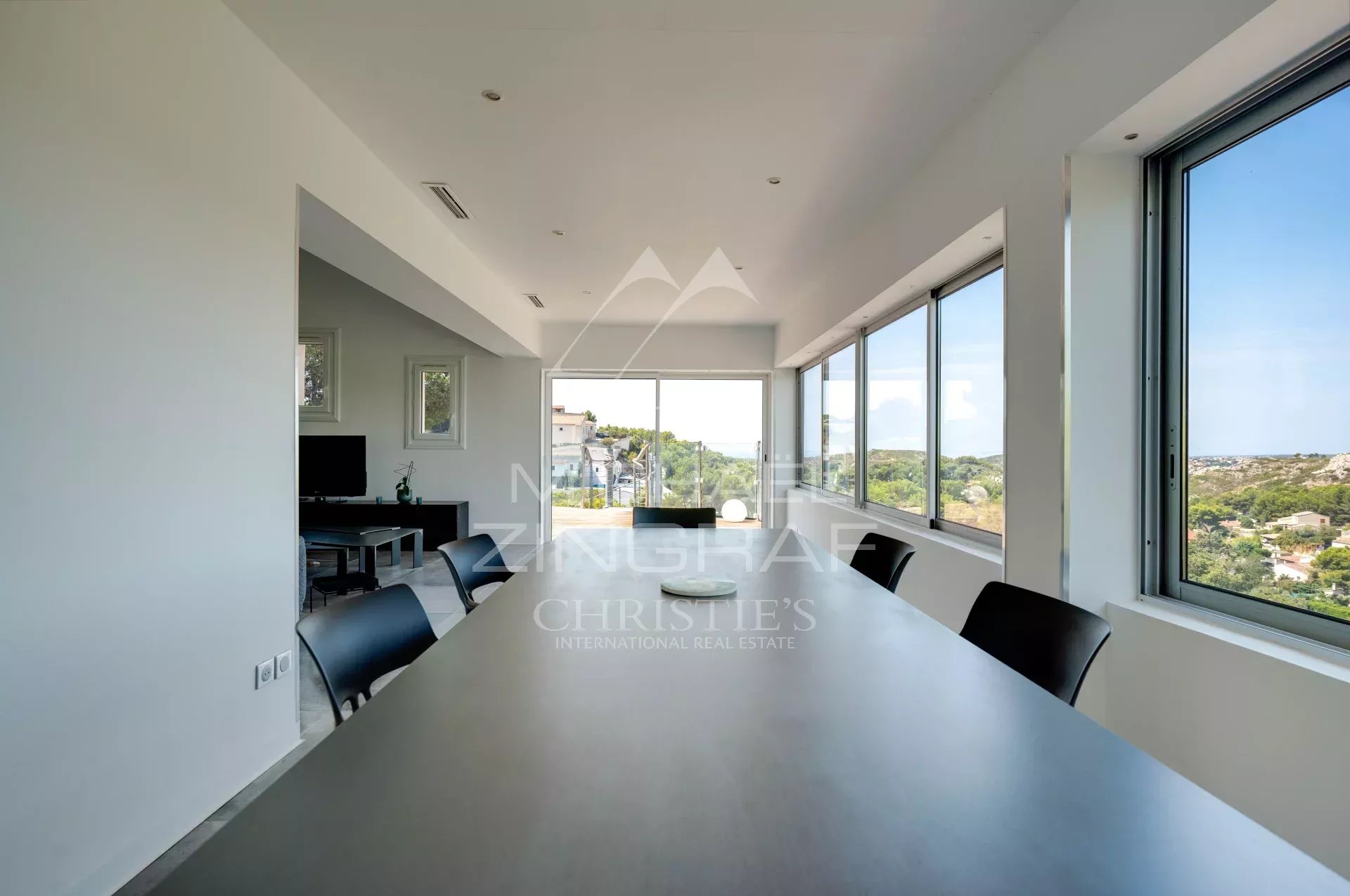 Intérieur d'une maison moderne avec vue sur la mer, grandes fenêtres et table à manger.