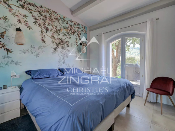 Chambre avec lit bleu et mur décoré de motifs floraux, fenêtre donnant sur la nature.