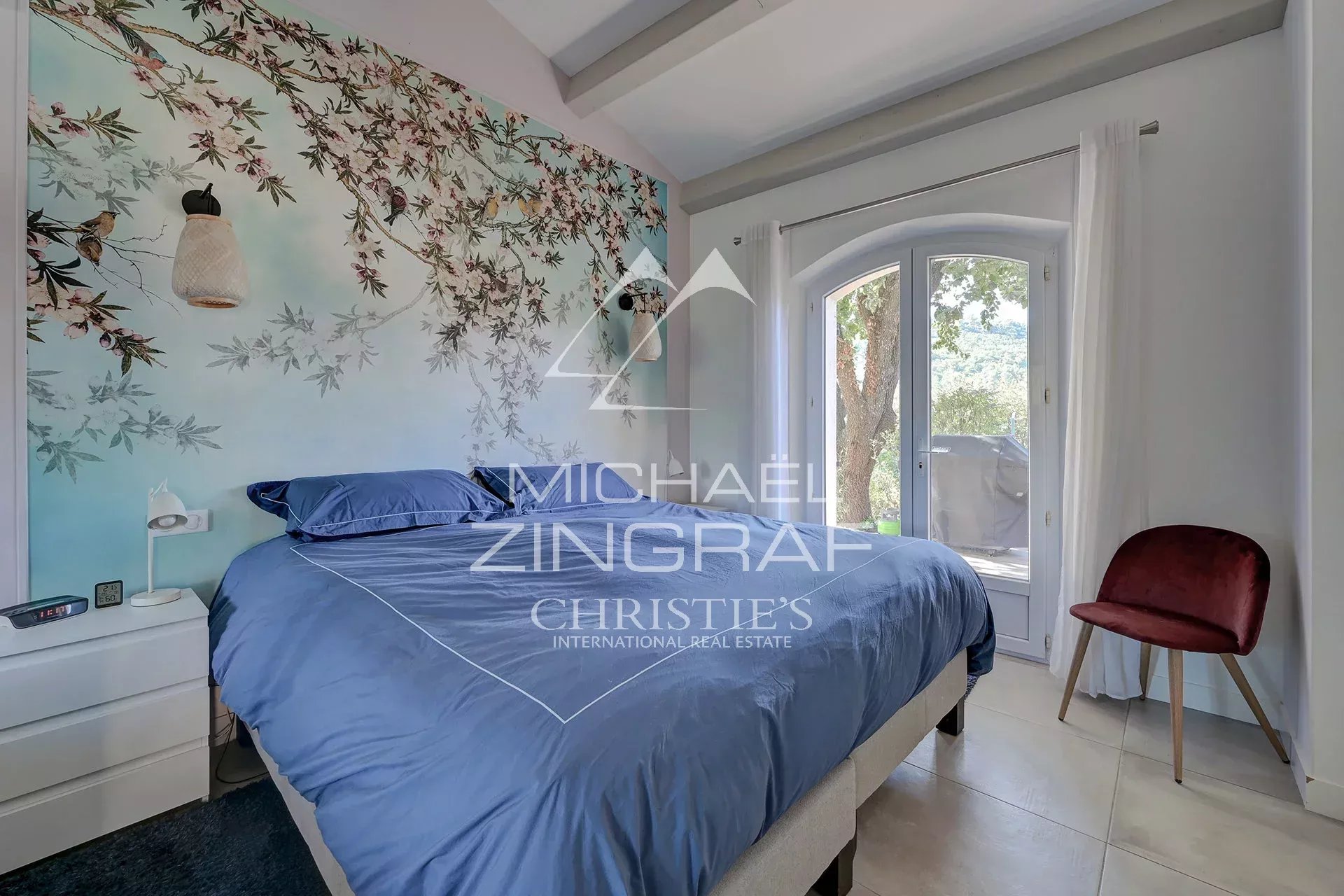 Chambre d’une propriété à Ansouis Chambre avec lit bleu et mur décoré de motifs floraux, fenêtre donnant sur la nature.