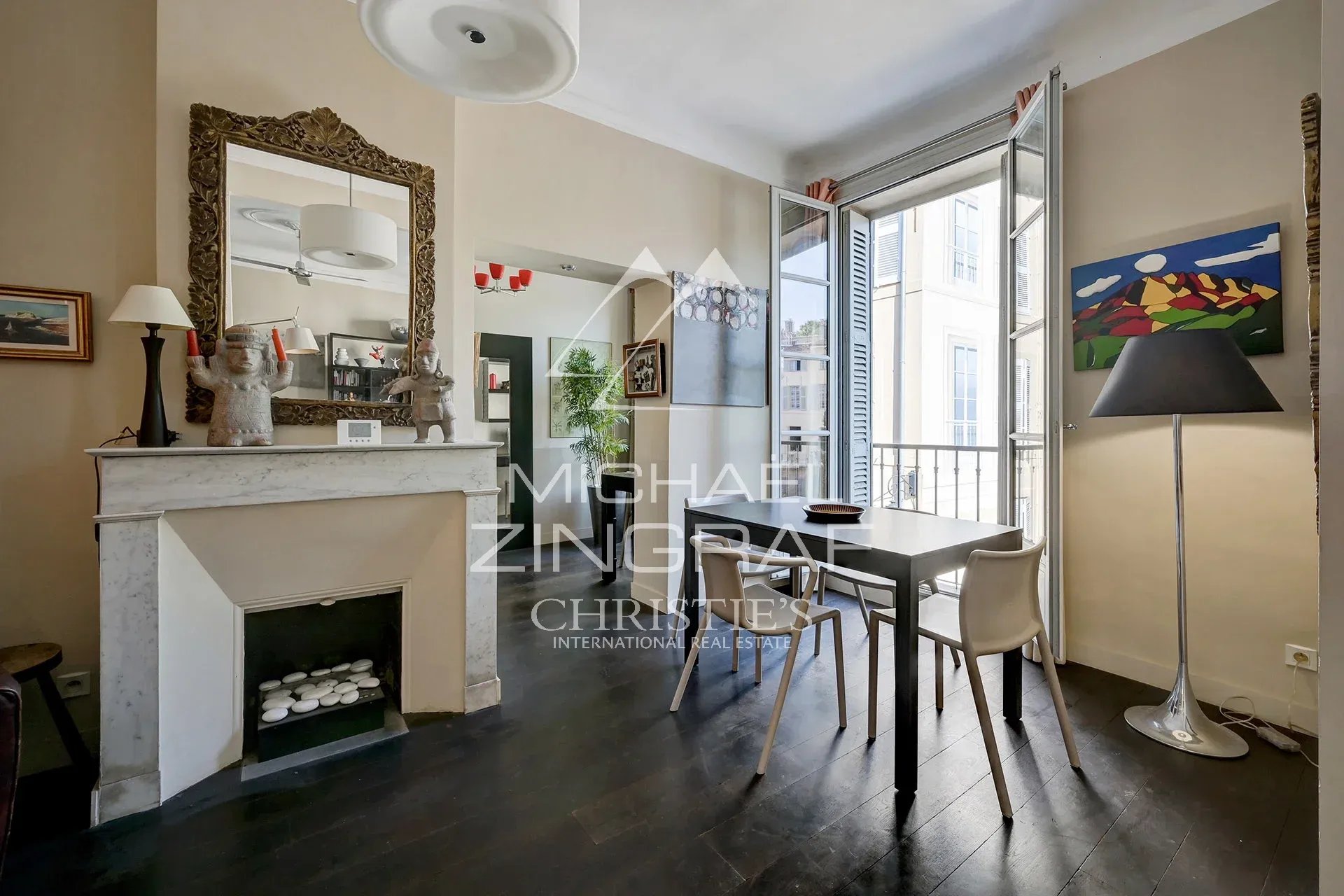 Intérieur d'un appartement ancien avec cheminée, table et fenêtres ouvertes sur la place des Prêcheurs à Aix-en-Provence.