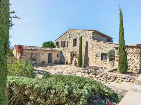 Stone farmhouse courtyard with a metal table and chairs, cypress trees, and blue sky above.