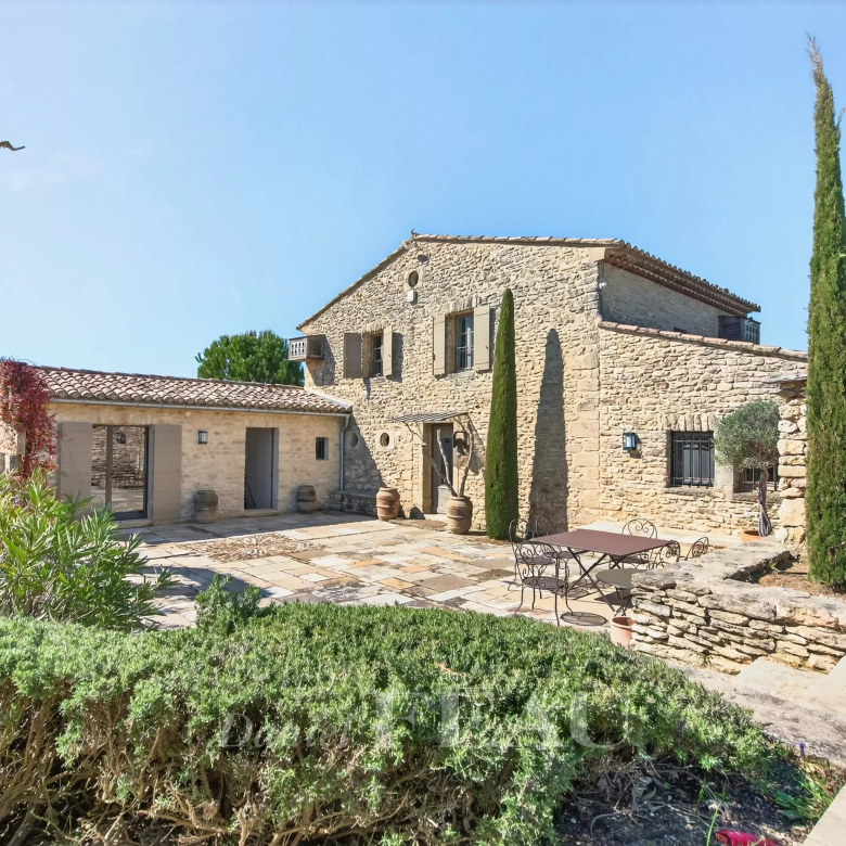 Stone farmhouse courtyard with a metal table and chairs, cypress trees, and blue sky above.