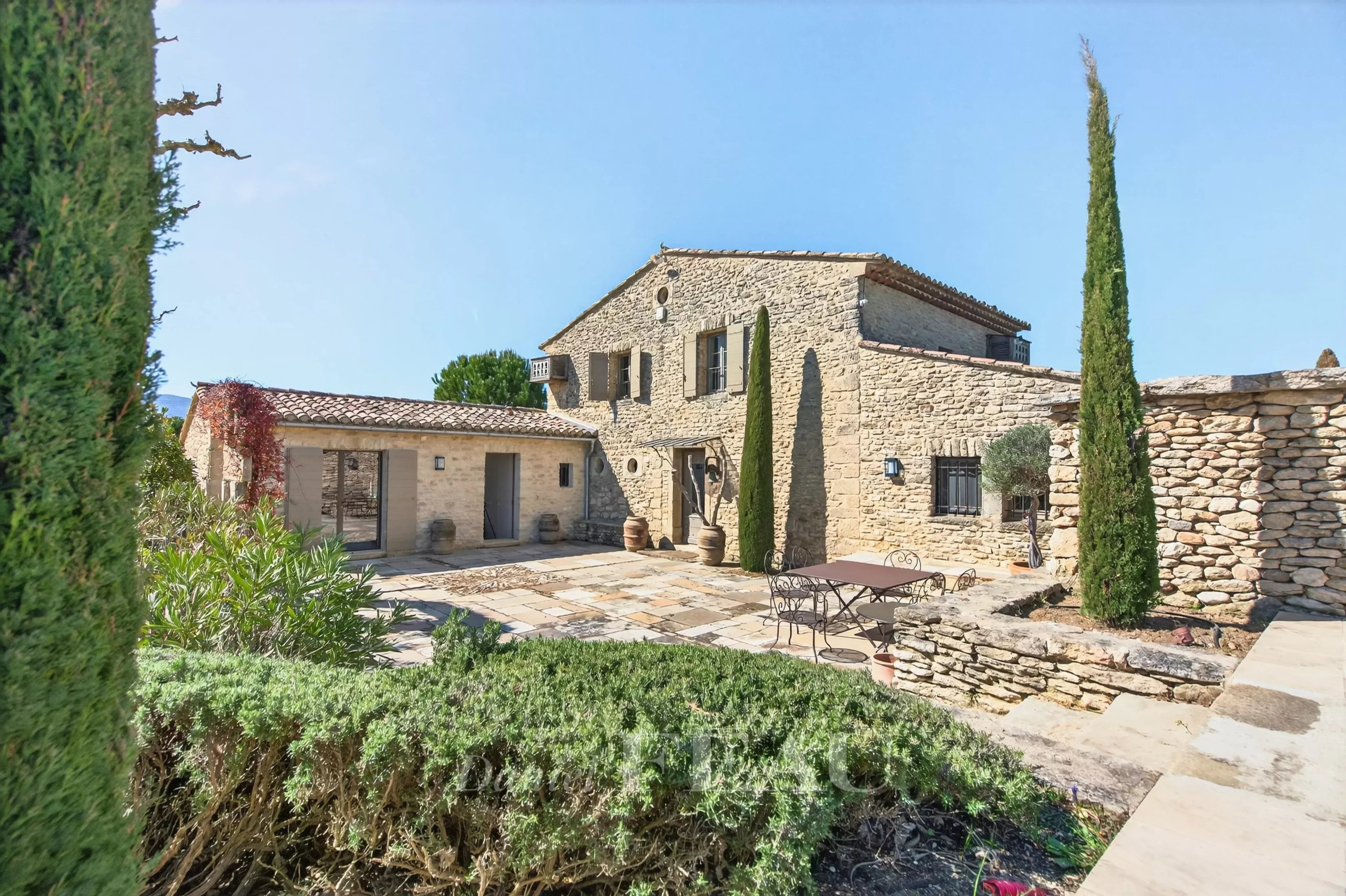 Stone farmhouse courtyard with a metal table and chairs, cypress trees, and blue sky above.