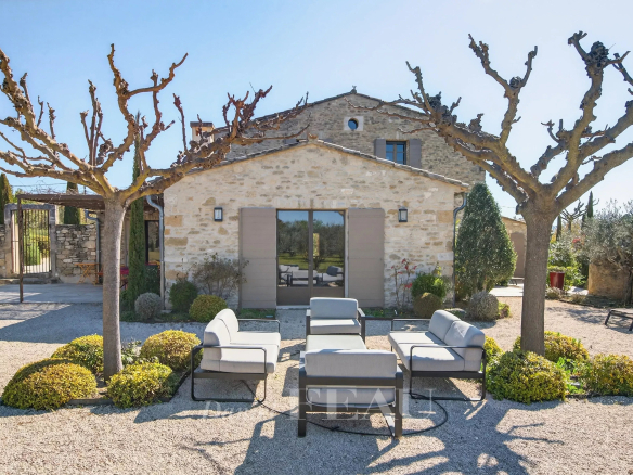 Stone cottage with trimmed trees and a gravel courtyard featuring a gray outdoor seating set of sofas and chairs arranged around a central table patio area