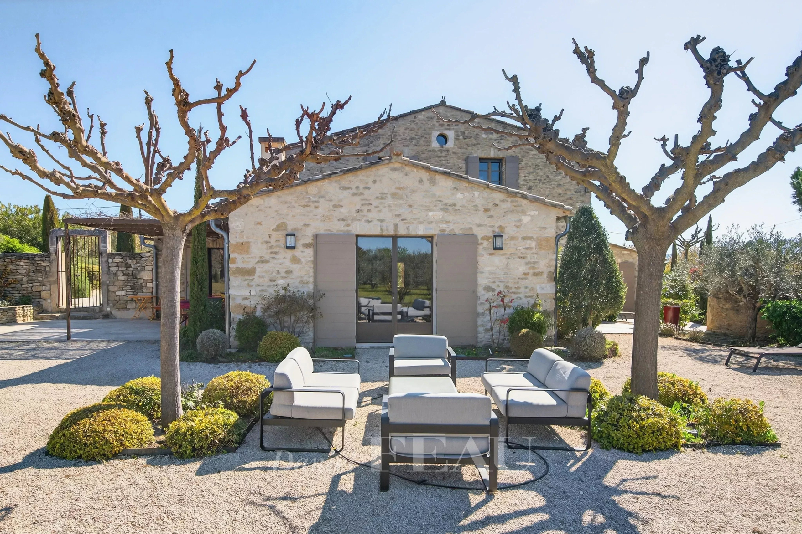 Stone cottage with trimmed trees and a gravel courtyard featuring a gray outdoor seating set of sofas and chairs arranged around a central table patio area