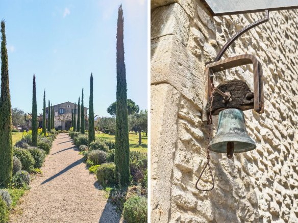 Path lined with tall cypress trees leading to a stone house in a sunny garden. A gravel walkway and manicured shrubs frame the scene.