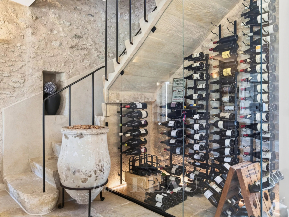 Wine cellar with floor-to-ceiling glass wall showing stacked wine bottles on metal racks beside a stone staircase and a large antique pot in the foreground.