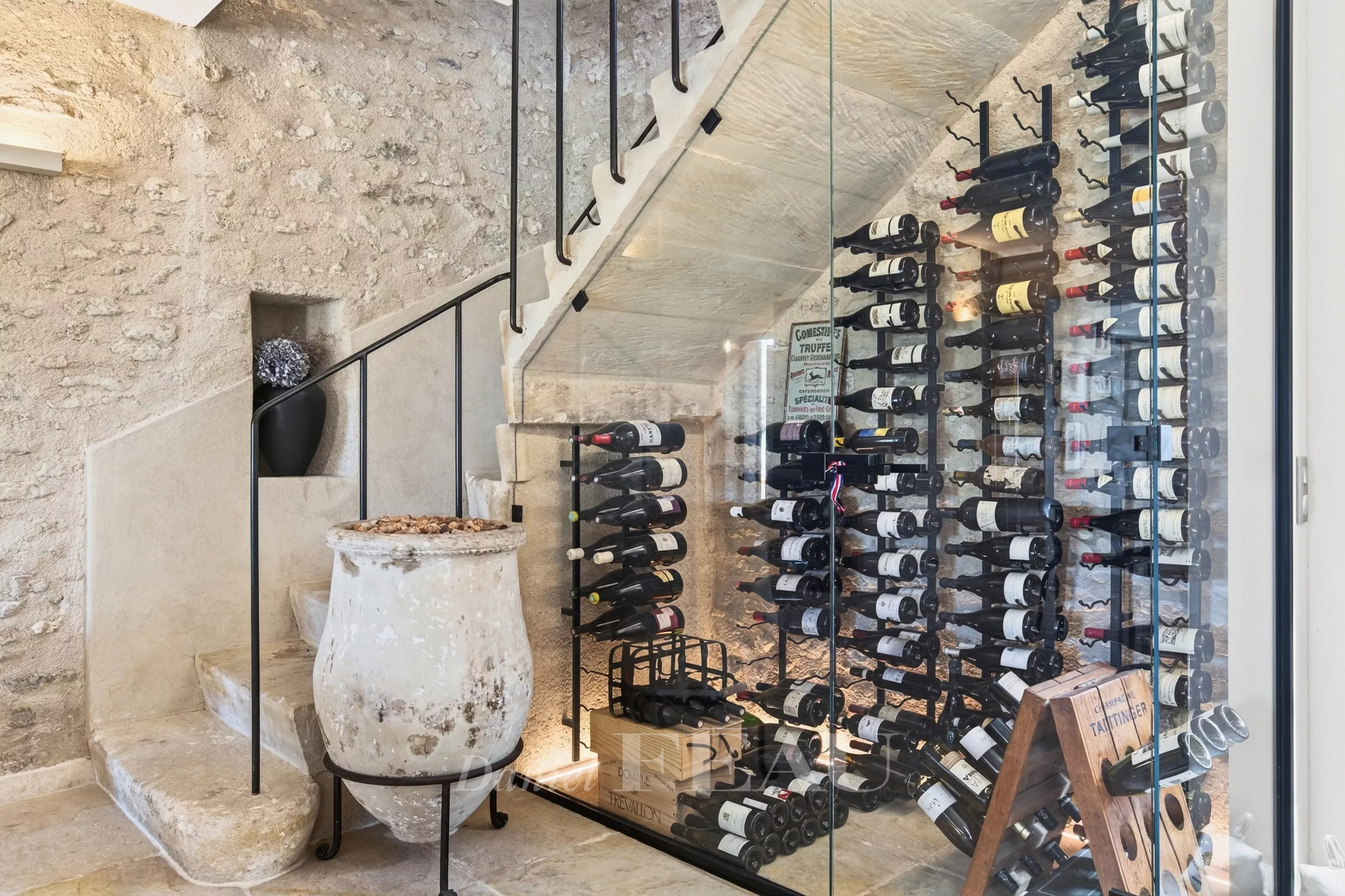 Wine cellar with floor-to-ceiling glass wall showing stacked wine bottles on metal racks beside a stone staircase and a large antique pot in the foreground.