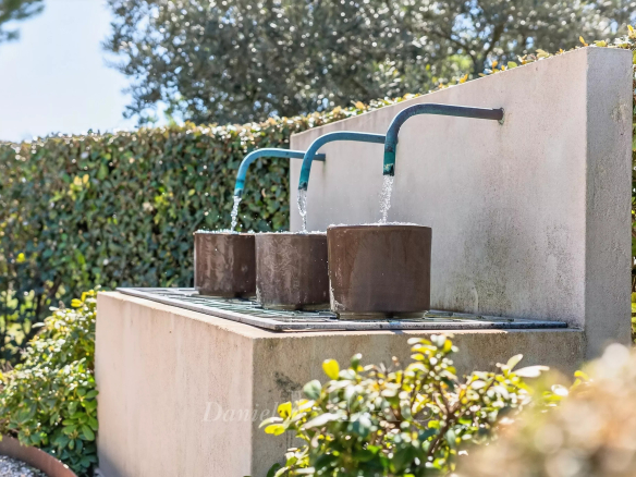 Outdoor drinking fountain with three blue taps dispensing water into brown cylindrical basins set on a concrete ledge with greenery in the background.