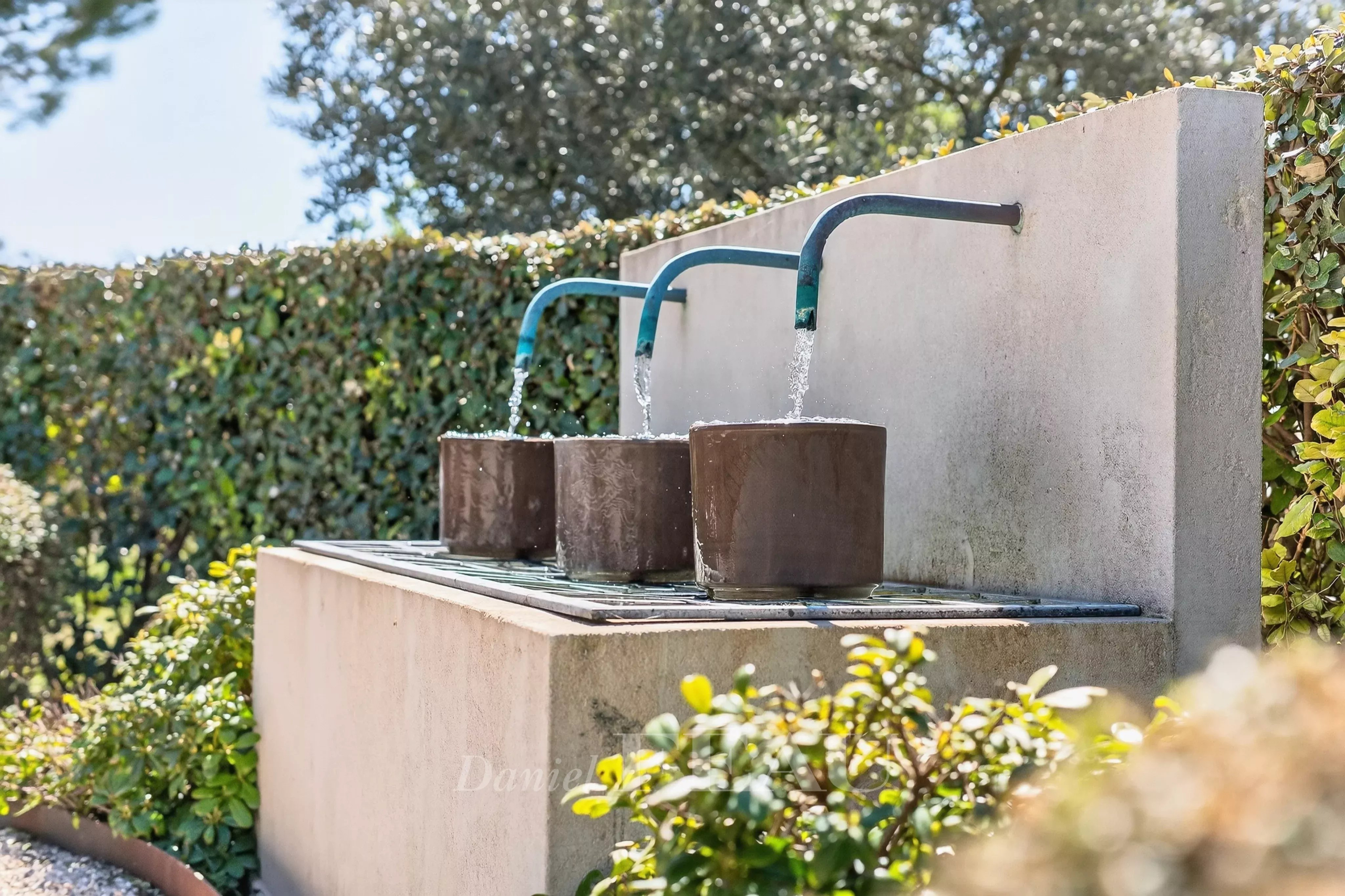 Outdoor drinking fountain with three blue taps dispensing water into brown cylindrical basins set on a concrete ledge with greenery in the background.