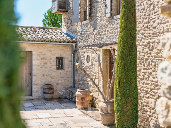 Sunlit stone courtyard with a tall green cypress, clay barrels, and rustic doors set in old stone walls.