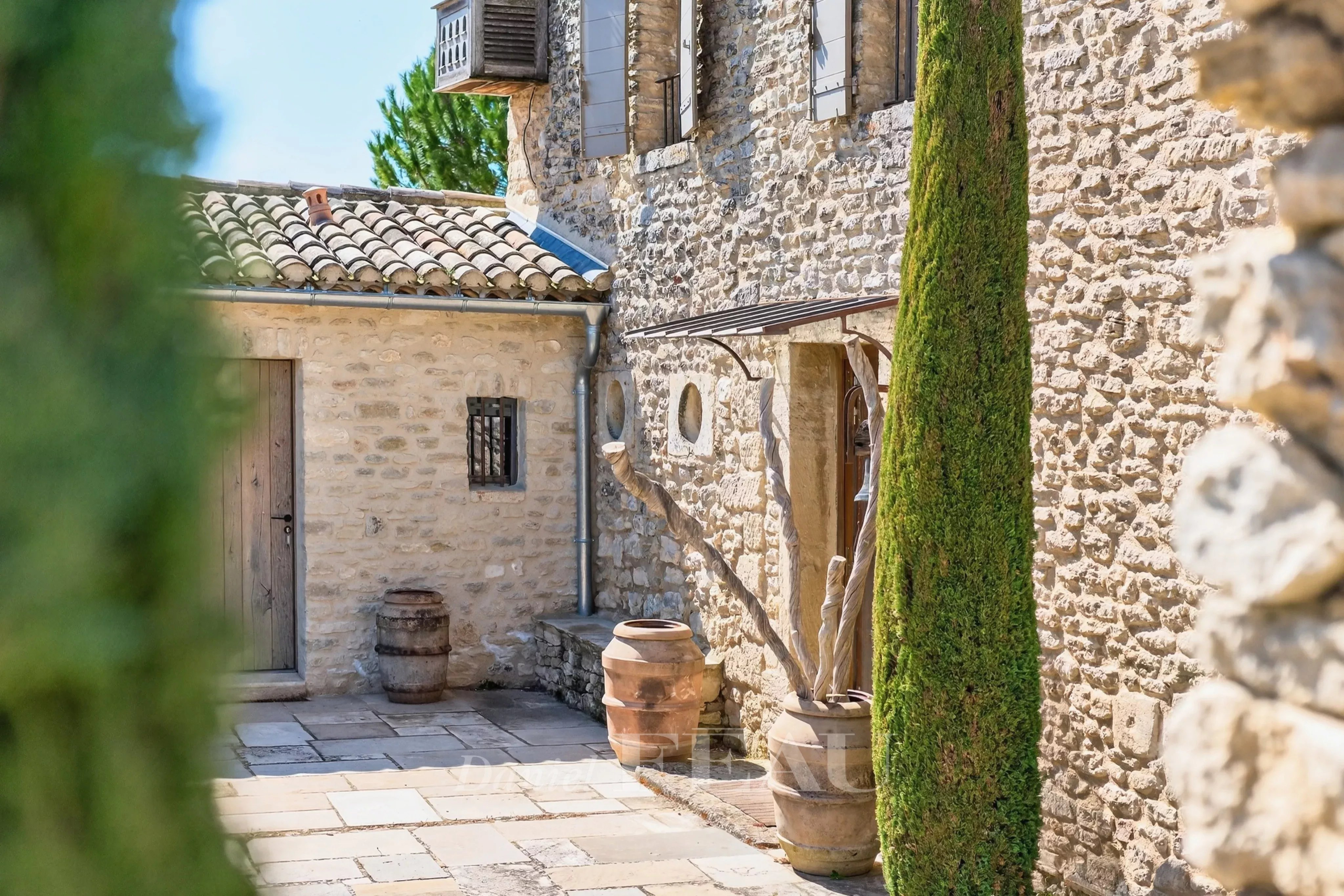 Sunlit stone courtyard with a tall green cypress, clay barrels, and rustic doors set in old stone walls.
