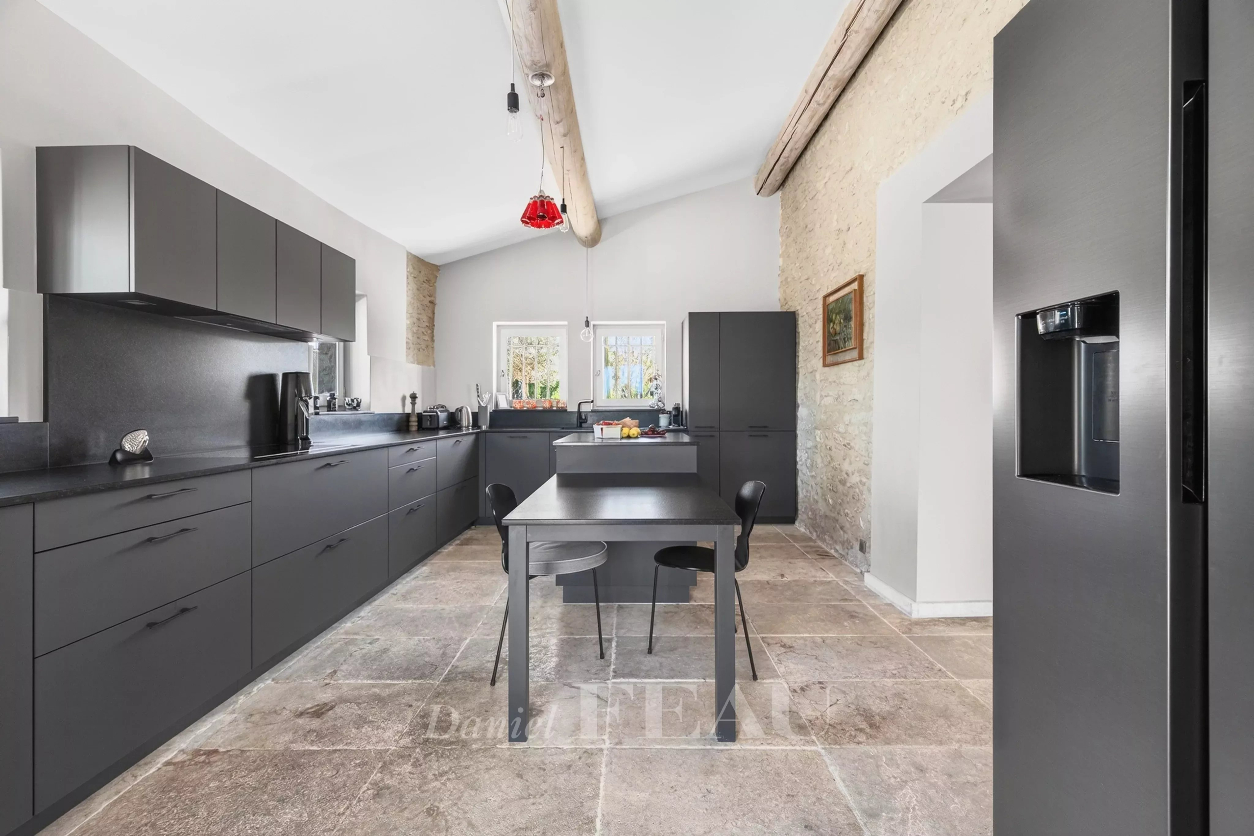 Modern grey kitchen with dark cabinets, stone wall, and a central island with two chairs under wooden ceiling beams.