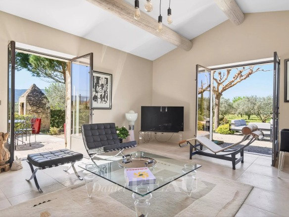 Bright living room with beige walls, exposed wooden beams, and a glass coffee table flanked by black leather chairs, open doors to a sunny garden.