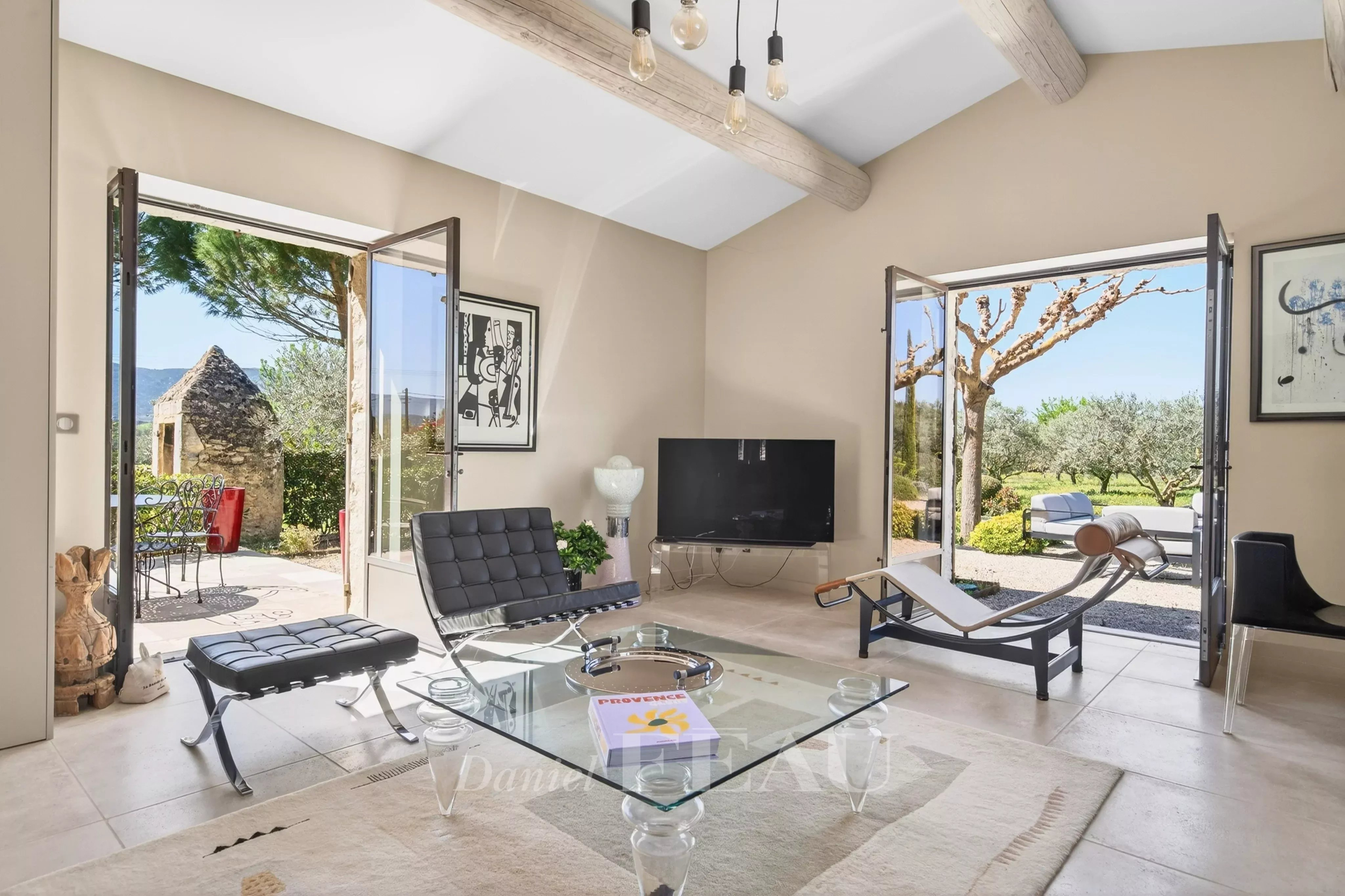 Bright living room with beige walls, exposed wooden beams, and a glass coffee table flanked by black leather chairs, open doors to a sunny garden.