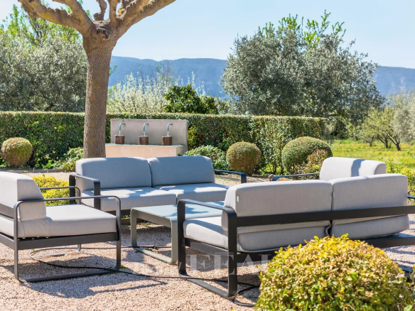 Outdoor seating area with modern gray-cushioned sofas and a glass coffee table on a gravel patio under a tree, garden and hills in the background