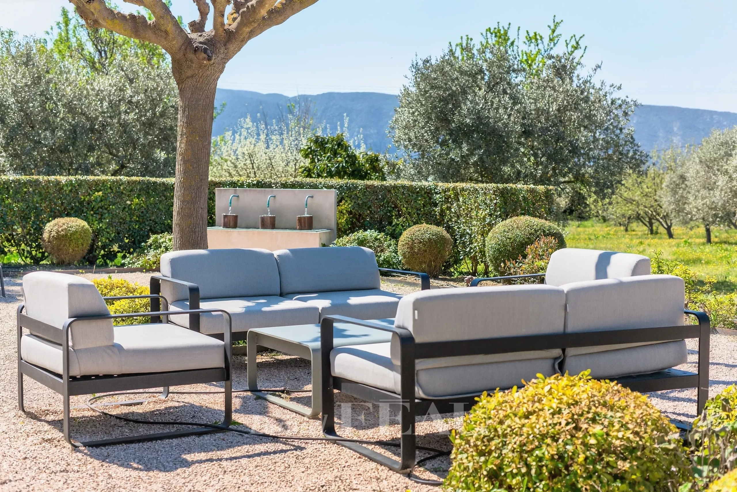 Outdoor seating area with modern gray-cushioned sofas and a glass coffee table on a gravel patio under a tree, garden and hills in the background