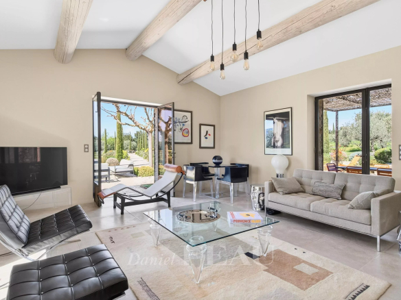 Bright open-plan living room with beige walls, exposed wooden ceiling beams, a glass coffee table, gray sofa, black leather lounge chairs, and large glass doors to a sunny patio.