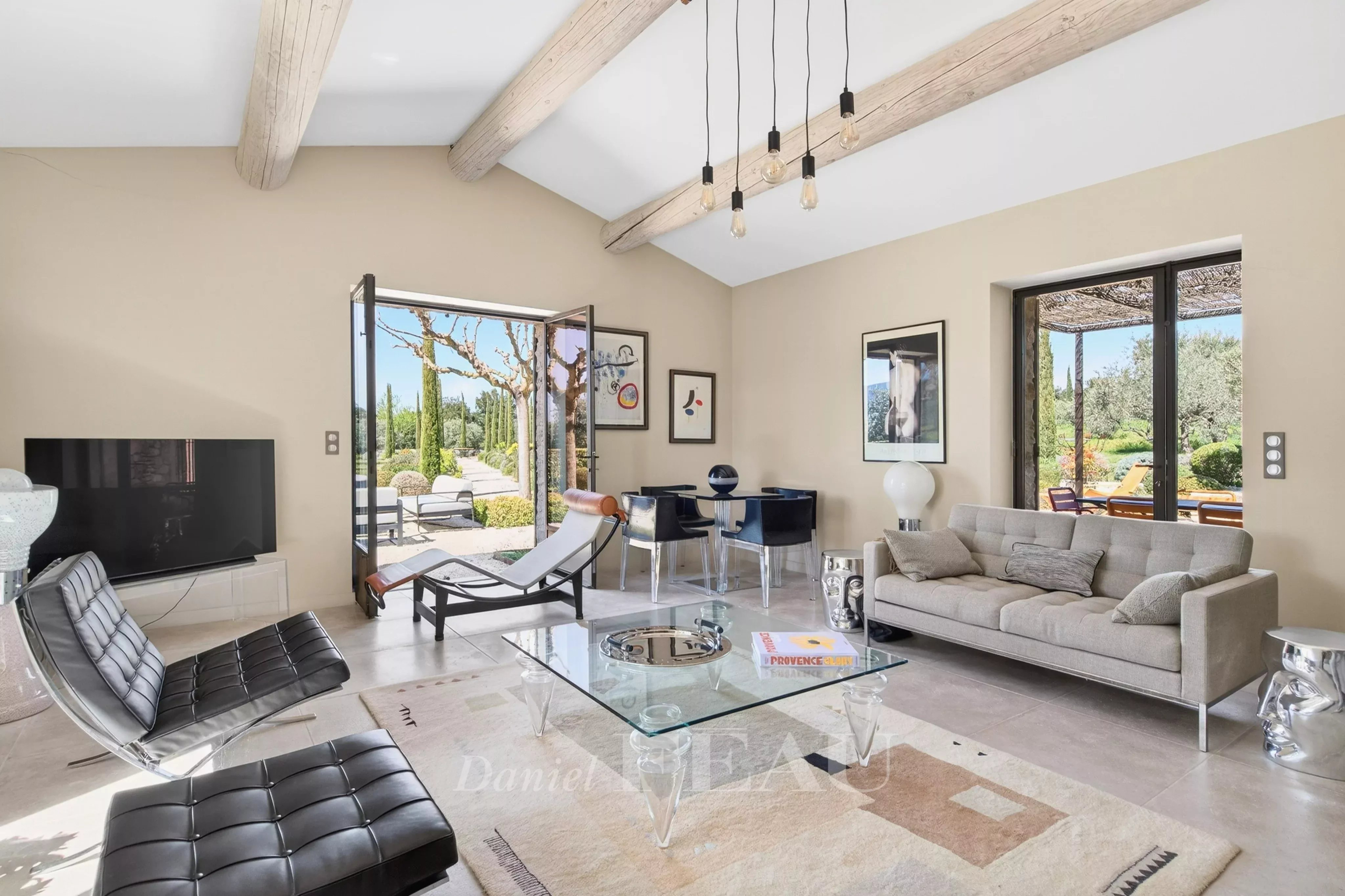 Bright open-plan living room with beige walls, exposed wooden ceiling beams, a glass coffee table, gray sofa, black leather lounge chairs, and large glass doors to a sunny patio.