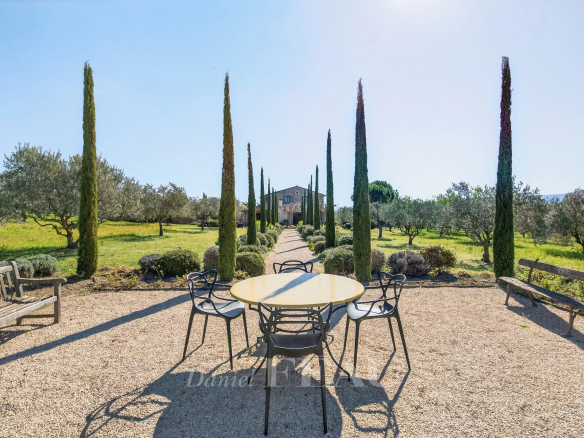 Outdoor garden scene with a round yellow table and black chairs on gravel, lined by tall cypress trees leading to a stone house in the distance; benches flank the area.