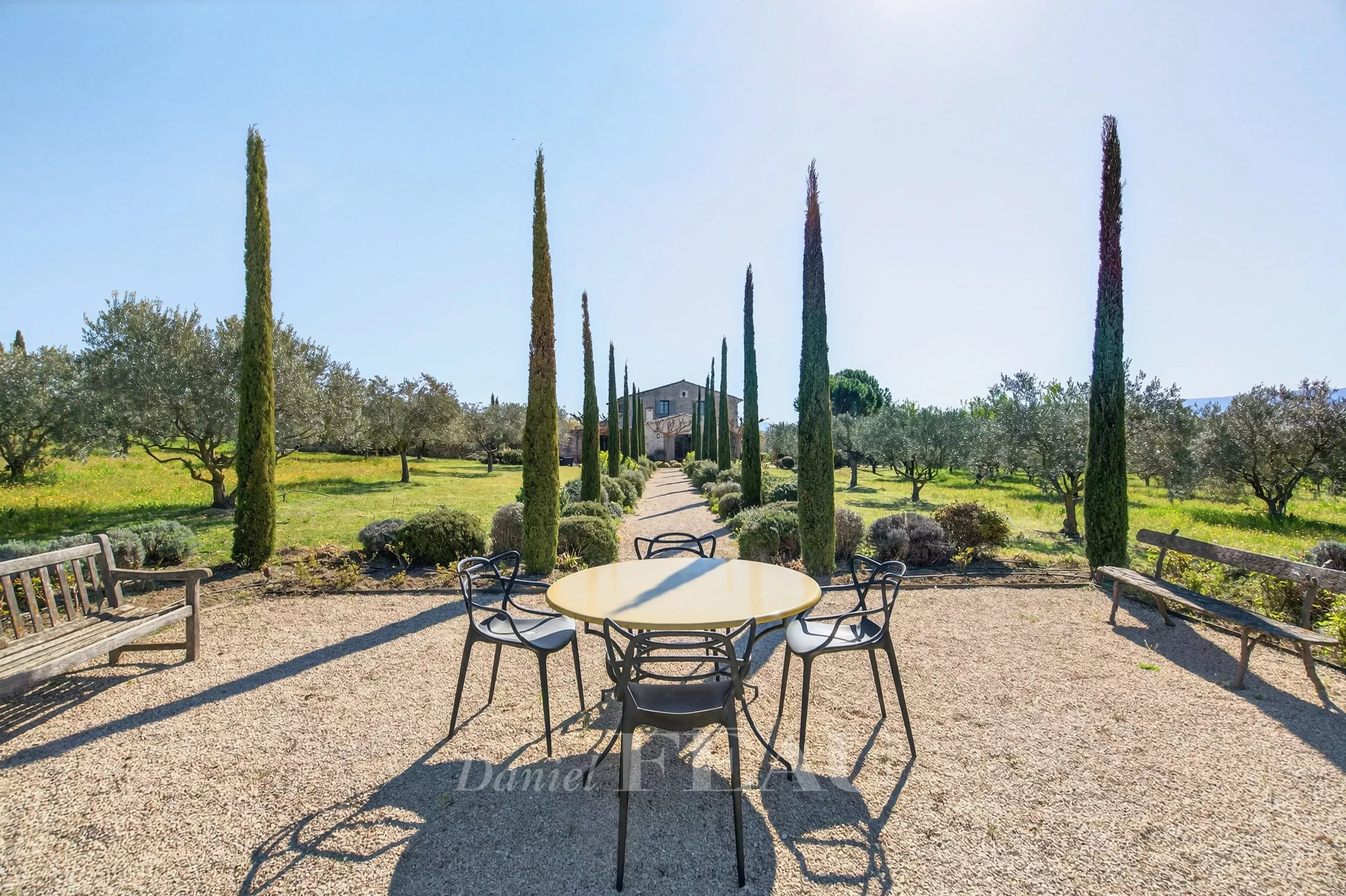 Outdoor garden scene with a round yellow table and black chairs on gravel, lined by tall cypress trees leading to a stone house in the distance; benches flank the area.
