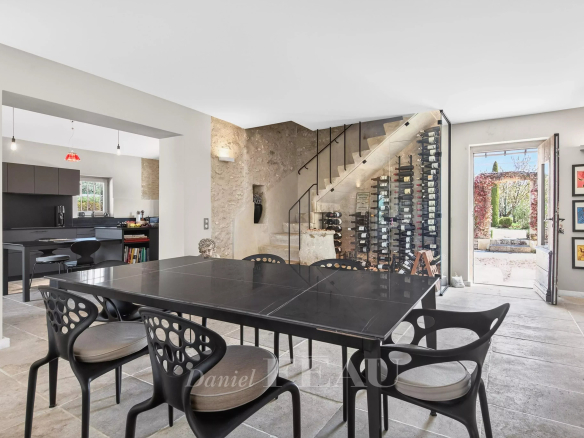 Open-plan dining room with a large black table, modern perforated chairs, and a tall glass-wine wall beside a stone staircase area; kitchen area visible in the background.