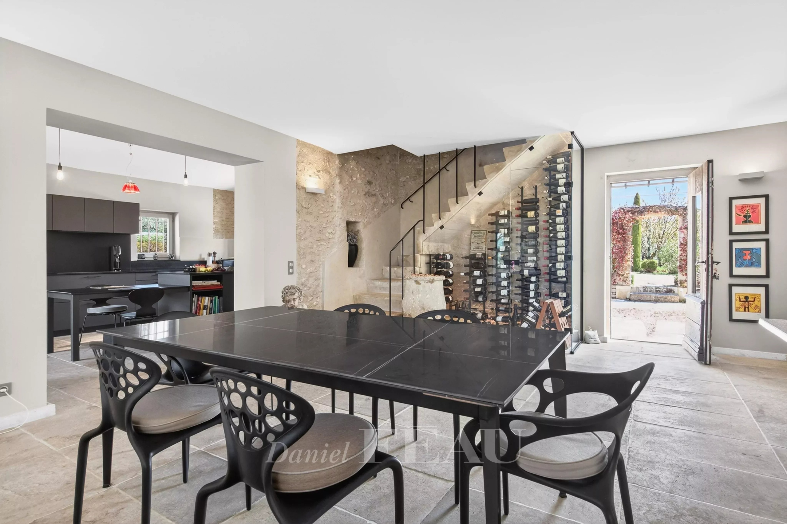 Open-plan dining room with a large black table, modern perforated chairs, and a tall glass-wine wall beside a stone staircase area; kitchen area visible in the background.
