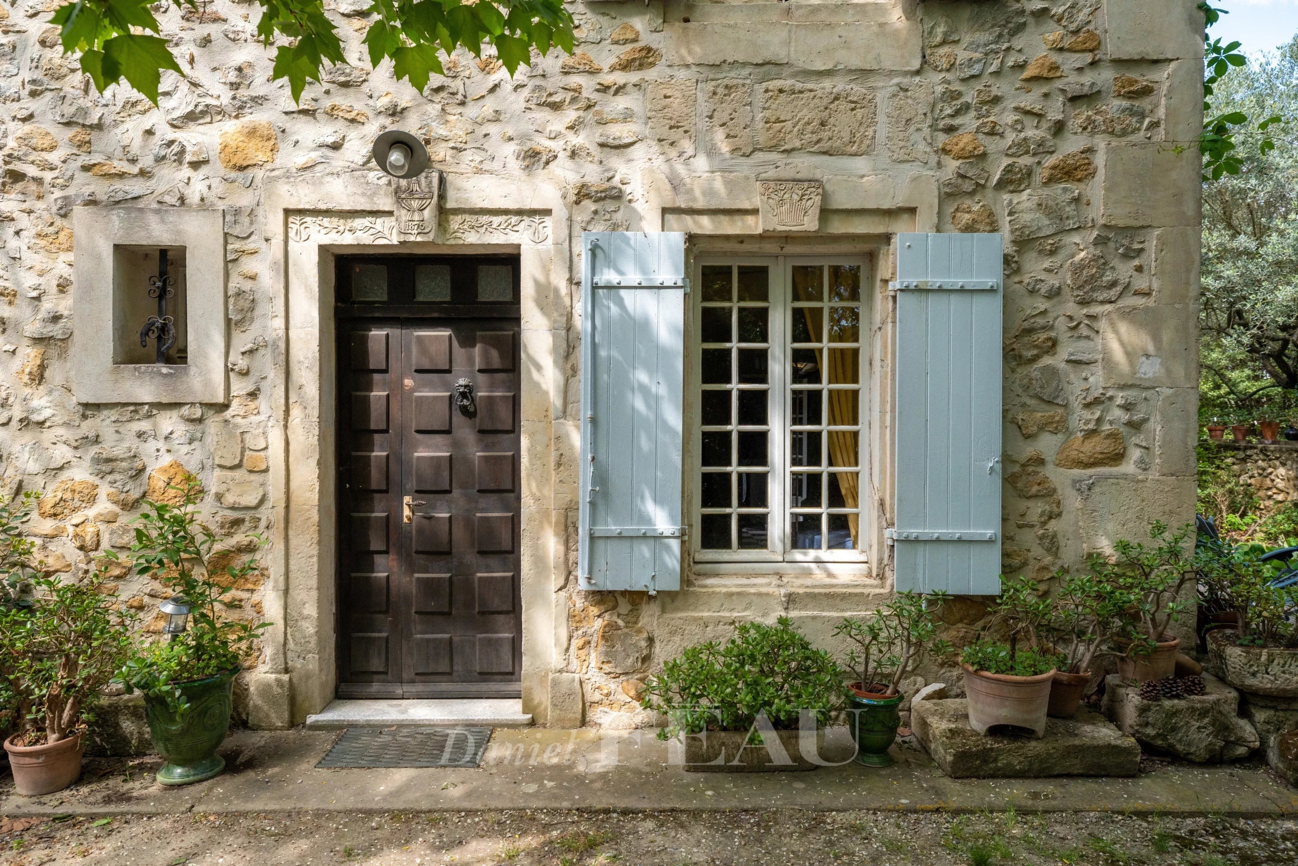Entrée d'un mas en pierre avec porte en bois et fenêtres à volets bleus, entouré de verdure.