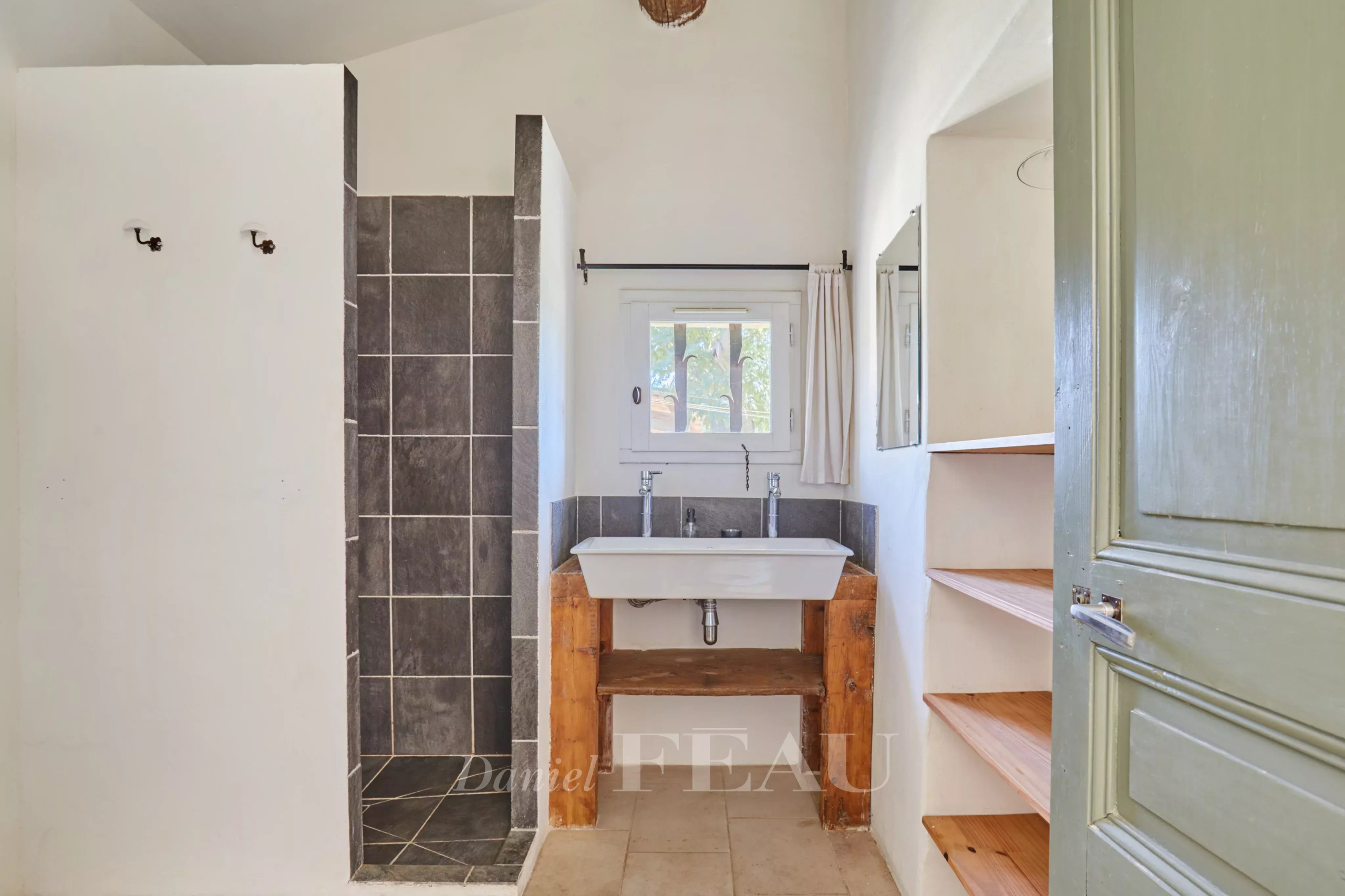 Salle de bain avec douche, lavabo en bois et étagères, murs blancs et carrelage gris.
